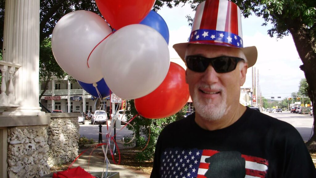 A man wearing patriotic hat and sunglasses with balloons.